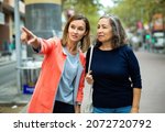 Small photo of Polite young woman pointing way to aged female tourist on city street on warm autumn day.