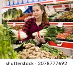 Small photo of Positive girl who works as a salesperson in a store, puts bundles of precocious chinese cabbage on the counter