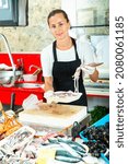 Small photo of Portrait of glad cheerful smiling saleswoman of seafood store proffering fresh squids for sale