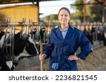 Small photo of Portrait of positive experienced female farmer standing with pitchfork near stall with dairy cows of Holstein breed in livestock farm