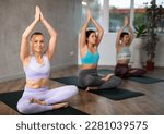 Small photo of Sedulous women doing lotus pose of yoga on black mat in light gym room with pot plants