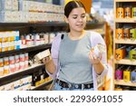 Portrait of interested young woman carefully reading labels on tin cans with choosing right hypoallergenic food for her dog in pet store