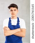 Small photo of Positive male worker in blue overall poses with folded arms on chest on gray background. Guy roustabout crossed his arms
