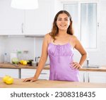 Small photo of Portrait of positive attractive girl in nightdress standing at kitchen table at home.