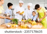 Small photo of Amiable woman and young man, skilled chefs wearing white cook jackets and toques, running culinary courses for preteen children, sharing secrets of cooking