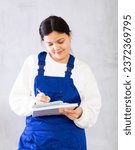 Small photo of Positive amiable young female cleaning service worker in blue uniform holding workbooks in hands and noting details of order. Studio portrait on gray background..