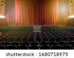 Large illuminated theatre hall with empty seats viewed from the rear looking towards the stage with its closed red curtain in a performing arts concept