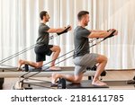 Full body side view of strong men in sportswear doing hug tree kneeling pilates exercise with resistance bands on reformer bed during training in gym