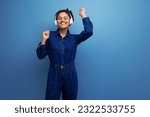 young joyful brunette latin woman dressed in blue denim overalls listens to music in wireless headphones on a blue background