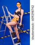Small photo of Isolated on blue, attractive brunette caucasian builder girl in denim shorts, black snickers, builder belt, black bikini and yellow build glasses stand on metal ladder and hold nail gun down