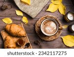 Small photo of Autumn aesthetic concept. Cup of cappuccino coffee with croissants, candles, plaid and yellow autumn leaves on wooden background close up.