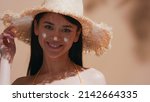 Small photo of Young brown-haired asian woman in a straw hat applies some sunscreen on her nose, enjoys the sun and smiles for the camera against beige background | Sunscreen commercial