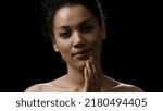 Small photo of Beautiful slim African American young black-haired woman touches her face looking at the camera and smiling on black background | Sagging jowls prevention concept