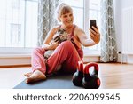 Small photo of Middle-aged, curvaceous, blonde woman with hair tied in bun, sits on gymnastic mat next to dumbbells, takes selfie for memory with beloved cat. Relaxing yoga exercises with pets for overweight adult