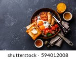 English Breakfast in cooking pan with fried eggs, sausages, bacon, beans, toasts and coffee on dark stone background copy space