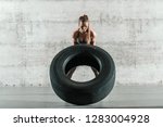 Strong dedicated female bodybuilder flipping tyre in crossfit gym. In background wall. Full length.