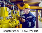 Serious caucasian unshaven worker in protective uniform and with hardhat using tablet for checking temperature in pipes. Factory interior.