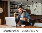 A happy businessman is sitting in a cafe, drinking coffee and watching a webinar on the laptop. The man has earphones in his ears so he can listen to the lecture. A businessman using laptop in cafe