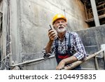 Small photo of An old factory worker with a helmet on his head, leans on the railing inside of the factory and communicates with his coworkers over the walkie-talkie. Workers talking with walkie-talkie.