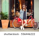 Cute girl in a summer dress, denim jacket, sunglasses stands with red vintage bicycle in a European city. Sunny summer. On background surf cafe.
