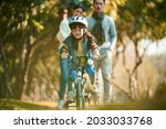 Small photo of little asian girl with helmet and full protection gears riding bike in city park with parents watching from behind