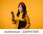 Small photo of Against a sunny yellow background, young Asian woman adeptly balances her laptop and smartphone, donning a yellow sweater and red beret.