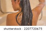 Small photo of CLOSE UP: Back view shot of beautiful woman rinsing her hair with shower handle. Pretty young lady showering her dark long hair while sitting in bathtub. Time for personal hygiene in home bathroom.