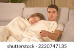 Small photo of CLOSE UP: Young couple resting under blanket and watching TV while catching flu. Twosome having a seasonal cold and snuggling on cosy sofa. Man watching a movie while woman is being tired and sleeping