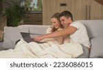 Small photo of CLOSE UP: Cheerful married couple sitting on couch and watching photos on laptop. Loving couple sitting on couch under blanket looking at laptop screen. Twosome enjoying at home on a cold winter day.