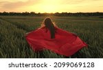 Small photo of Happy child girl plays superheroes, she runs across the green field in a red cloak, the cloak flutters in the wind. The concept of children's play, dreams. Young super girl hero outdoors