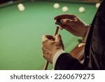 Close up of man in a jacket rubs the end of a cue with chalk before the game. Background of green pool table with billiard balls