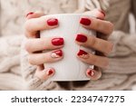 Woman's hands with red manicure and gold foil on the nails. Trendy winter nail design. Woman with a beautiful manicure holding a white knitted cup. The concept of cozy Christmas holidays and New Year.
