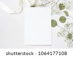 Styled stock photo. Feminine wedding desktop stationery mockup with blank greeting card, baby's breath Gypsophila flowers, dry green eucalyptus leaves, satin ribbon and white background. Empty space. 