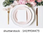 Feminine wedding, birthday desktop mock-up scene. Porcelain plates, blank paper greeting, menu card, ribbon, golden cutlery, leaves,roses, peony flowers. White table background. Flat lay, top view