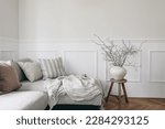 Stylish living room with vase and blooming cherry plum tree branches. Springtime home decor. Elegant scandinavian interior with comfy sofa, linen cushions and blanket. White wall background. 