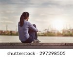 Rear View of a Thoughtful Young Woman Facing at the River while Sitting on a Long Concrete Bench and Hugging her Legs