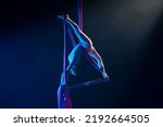 Small photo of Female circus gymnast hanging upside down on aerial silk and demonstrates stretching. Young woman performs tricks at height on silk fabric. Acrobatic stunts on black background with blue backlight.