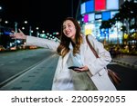 Small photo of Asian business woman walking to hail waving hand taxi on road in city street at night, Beautiful woman smiling using smartphone application hailing with hand up calling cab outdoor after late work
