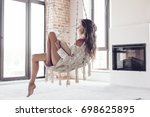 Young woman chilling at home in comfortable hanging chair near fireplace. Girl relaxing and reading book in swing in loft living room with brick walls.