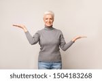 Small photo of Make right choice. Smiling aged woman comparing variants, making scales with her empty hands over light studio background with copy space
