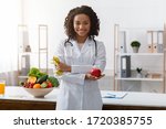 Small photo of African female dietician with crossed arms holding fresh apple, posing at workplace