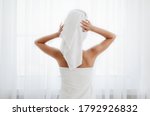 Back view of woman wrapped in towel standing next to window, drying her hair after morning shower