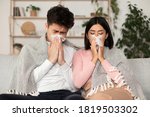 Small photo of Ill Asian Couple Having Cold, Blowing Nose Sneezing In Paper Tissue Sitting On Couch At Home Feeling Unwell. Rhinitis And Seasonal Allergy, Flu Illness. Family On Sick Leave Self-Isolation Concept
