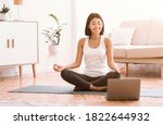 Keep Calm. Happy asian woman meditating with trainer online via laptop connection, copy space. Healthy lady sitting on the floor on yoga mat in lotus position, looking at computer and smiling