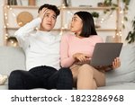 Small photo of Asian Shopaholic Wife Shopping Online With Shocked Husband, Asking Him Buy Expensive Clothes And Spend Too Much Money Sitting On Sofa At Home. Prodigality, Shopaholism And Bad Spending Habits Concept
