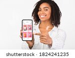 App For Online Store. Happy african woman holding and showing phone with clothes shop website and pointing at screen