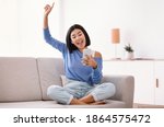 Chill And Lifestyle Concept. Exited asian woman wearing wireless earphones listening to favorite song, holding mobile phone, sitting on sofa and dancing in living room, raising hand and finger up