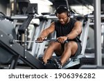 Motivated young black sportsman with wireless headset having workout on rowing machine. Handsome african american man bodybuilder training body on modern block exerciser in gym, copy space