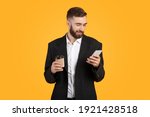 Positive young businessman checking email or browsing web on his cellphone, drinking takeaway coffee on orange studio background. Happy guy in formal wear using internet on mobile device