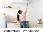 Back rear view of smiling woman selecting mode on cooking hood, standing near kitchen appliance in contemporary interior and decor with shelves, pushing button on mechanical fan above the stove